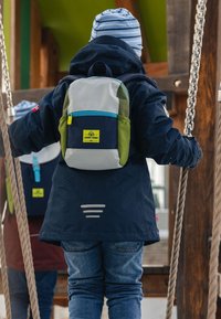 Enfant portant une veste bleu marine et un chapeau rayé, avec un petit sac à dos multicolore, escaladant des cordes sur un équipement de jeu en bois.