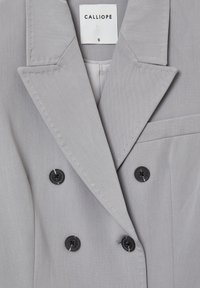 Blazer grigio doppiopetto con rever affilati, dotato di bottoni neri e taschino sul petto. Realizzato in tessuto morbido con una trama delicata.