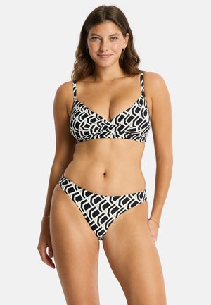 Kobieta z długimi brązowymi włosami nosi czarno-biały bikini z geometrycznym wzorem, stoi i uśmiecha się na tle białego tła.
