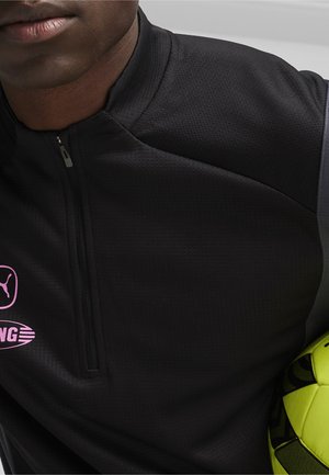 Schwarzes, mit Reißverschluss versehenes Sportoberteil aus strukturiertem Stoff, mit pinkfarbenen Logos von Puma und einer anderen Marke. Hält einen schwarz-neon-gelben Fußball.
