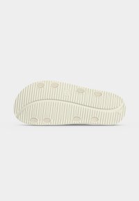 Semelle d'une chaussure blanche avec des rainures horizontales et sept coussinets ronds en caoutchouc beige sur un fond clair uni.
