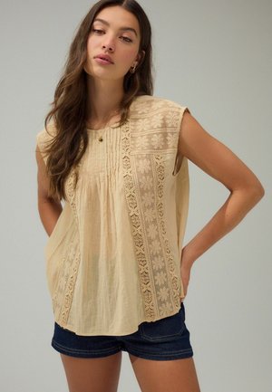 Light beige blouse with short sleeves, featuring embroidered lace details along the front and sides, with a loose and textured design.