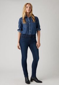 Tumma sininen farkkupaita napitettuna, rintataskut, yhdistettynä istuviin tummiin farkkuihin ja mustiin nilkkabootseihin.