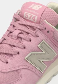 Adidași din suede roz cu un interior texturat, având un logo "N" gri, șireturi rotunde și o limbă căptușită care afișează numărul modelului 574.