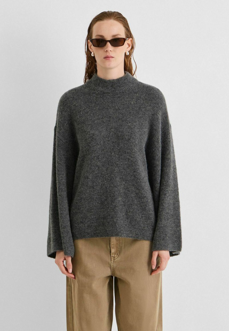 Femme portant un pull en maille gris foncé, un pantalon beige, des lunettes de soleil foncées et des boucles d'oreilles en perles, debout devant un fond clair uni.