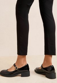 Črni lakasti usnjeni čevlji Mary Jane z okroglo konico, trakcem okoli gležnja in sijočim zaključkom. Nizka blok petka z rahlo teksturirano gumijasto podplatom.