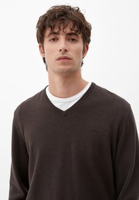 Brauner V-Ausschnitt-Pullover aus weichem Stoff, mit langen Ärmeln und geripptem Halsausschnitt. Getragen über einem weißen Rundhalsausschnittshirt, mit dezentem Markendetail.