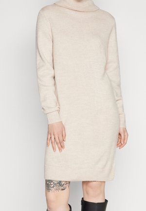 Kobieta ubrana w kremową dzianinową sukienkę z długim rękawem i dekoltem typu komin, czarne kozaki za kolano oraz z kwiatowym tatuażem na lewej udzie.