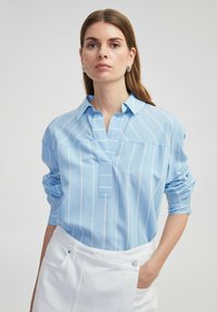Camisa de manga larga azul claro con rayas verticales blancas, con tapeta abotonada y cuello. Llevada con una falda blanca que presenta bolsillos laterales.