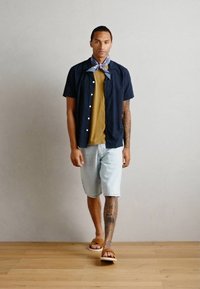 Chemise à manches courtes bleu marine sur un top moutarde, shorts en denim clair et sandales beige. Une écharpe bleue est nouée autour du cou.