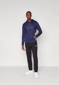 Sweatshirt com capuz azul-marinho com um grande logótipo, apresentando acentos laterais amarelos e azuis. Combinado com calças pretas e ténis brancos.