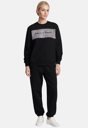 Mujer rubia con sudadera negra Maison Gaelle con panel de purpurina plateada y pantalones de chándal negros, de pie sobre fondo blanco.