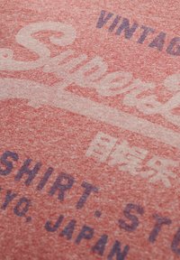 Camiseta de tejido rojo jaspeado con un gráfico de estilo vintage. Texto detallado en azul y blanco que incorpora caracteres japoneses. Textura suave.