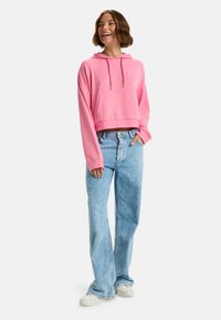 Różowa skrócona bluza z kapturem, z przednią kieszenią i sznurkiem, w połączeniu z jasnoniebieskimi szerokimi spodniami dżinsowymi i białymi trampkami.