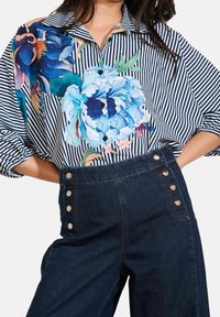 Femme portant une chemise rayée avec de grands motifs floraux bleus rentrée dans un pantalon en jean foncé taille haute avec des boutons dorés.