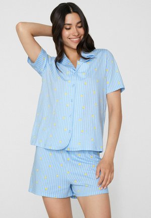 Lächelnde Frau mit langen dunklen Haaren, die ein hellblaues Hemd mit Knöpfen und passende Shorts mit gelbem Punktemuster trägt und mit einem erhobenen Arm posiert.