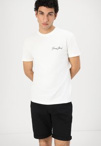 T-shirt en coton blanc à manches courtes, avec du texte brodé noir sur la poitrine gauche ; associé à un short noir retroussé.