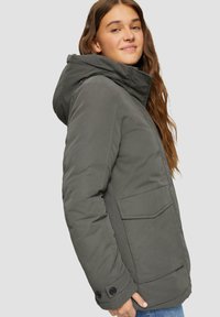 Olijfgroene geïsoleerde jas met een capuchon, voorzien van zijzakken, een voorflap en knopenaccenten op de mouwen. Gestructureerde stofafwerking.