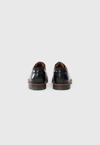 Un paio di scarpe eleganti in pelle nera con suole marroni, viste da dietro su uno sfondo bianco uniforme.
