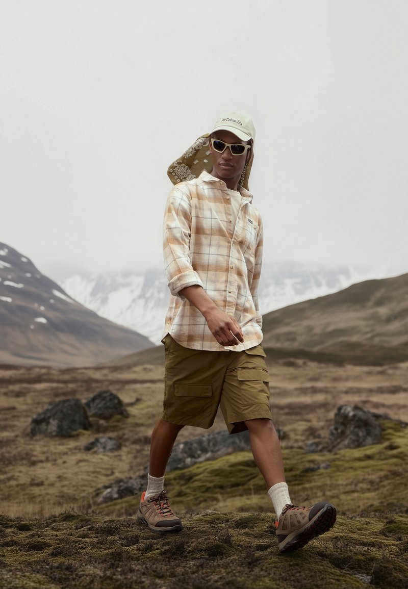 Camisa de cuadros beige de botones, pantalones cortos de cargo oliva y botas de senderismo marrones con detalles en naranja. Los accesorios incluyen gafas de sol y una gorra ligera.