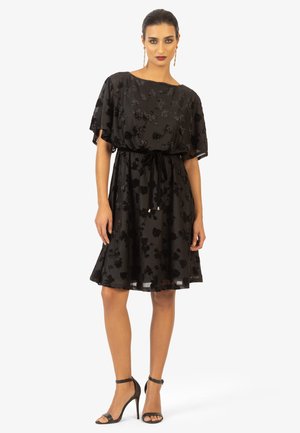 Schwarzes Kleid mit kurzen, lockeren Ärmeln und floralem Stickerei. Verfügt über einen Bindegürtel in der Taille und einen ausgestellten Rock. Getragen mit Riemchen-Heels.