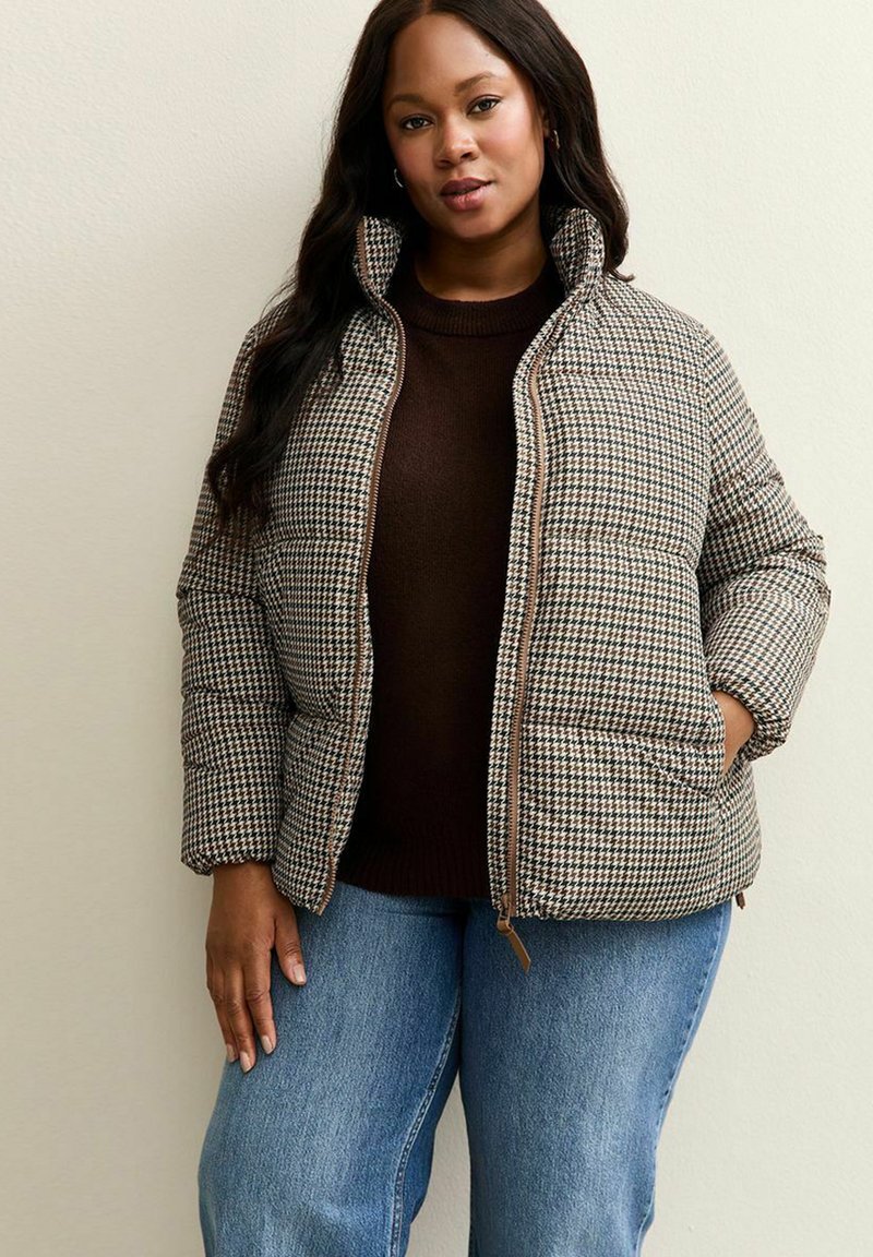 Giacca imbottita in un motivo houndstooth marrone e crema, con colletto alto, chiusura con zip e due tasche laterali. Indossata sopra un maglione marrone.