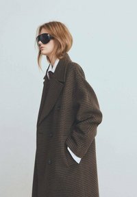 Abrigo largo de patrón pata de gallo marrón con diseño de doble botonadura, solapas grandes y mangas anchas; usado sobre una camisa blanca y corbata.