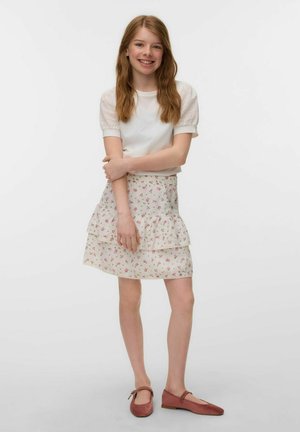 Uśmiechnięta dziewczyna z długimi jasnobrązowymi włosami, ubrana w biały top z bufiastymi rękawami, kwiecistą, warstwową spódnicę oraz brązowe buty typu Mary Jane, stojąca na jednolitym tle.