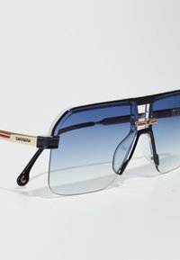 Ochelari de soare cu cadru negru, bara de sus clară și lentile cu un gradient albastru. Au detalii aurii pe brațe și un design unic cu suporturi pentru nas.