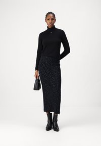 Melnā turtlenecks blūze, apvienota ar ceļgala garuma, raibumu melnu svārku un ceļgalu augstumā melnām zābakiem, tur rokā mazu melnu roku somiņu.