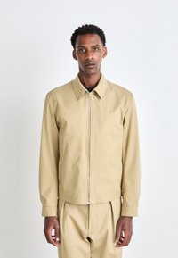 Chaqueta con cremallera color caqui y cuello puntiagudo, de mangas largas, fabricada en tejido suave. Presenta una cremallera plateada y un corte relajado.