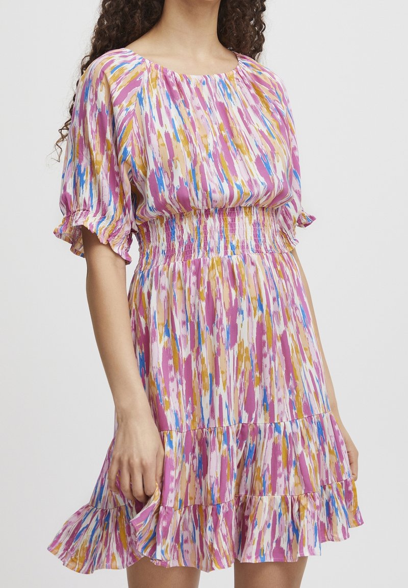 Femme portant une robe courte multicolore avec des motifs coups de pinceau rose, bleu et moutarde et des manches et un bas volantés.