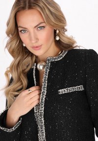 Jachetă din tweed negru cu decolteu rotund, margine ornamentată cu bețe și un mic buzunar pe piept. Prezintă un accent mare din perlă la guler.