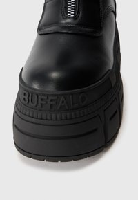Sapatilha plataforma preta com parte superior em couro liso, fecho com zíper e sola de borracha com detalhes texturizados e marca "BUFFALO".