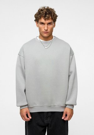 Grijze oversized sweatshirt van zachte stof, met een ronde halslijn, verlaagde schouders en geribbelde manchetten. Draagt het met zwarte broek.