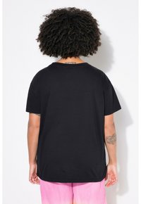 Sort kortærmet T-shirt med afslappet pasform, rund hals og struktureret stof; båret med lyserøde, plisserede bukser. Synlige tatoveringer på armene.