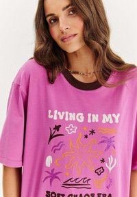 Př oversized růžové tričko s barevným grafickým designem, který zobrazuje slunce, květiny a vlny, a kontrastním hnědým žebrovým výstřihem.