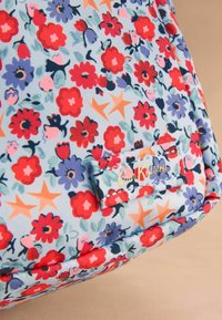 Ziedu raksts somā ar pasteļzilu fonu, kurā ir sarkani, rozā un violeti ziedi, akcentēti ar oranžām zvaigznēm. Iekļauta birka ar "Cath Kidston."
