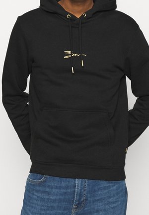 Homme portant un sweat à capuche noir avec un petit logo jaune et des cordons, associé à un jean bleu, mains dans le dos.