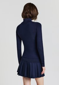 Frau mit schulterlangem braunem Haar trägt einen navyblauen langärmeligen Rollkragenpullover und einen gefalteten Minirock, steht vor einem hellen Hintergrund.