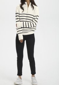 Suéter de punto color crema con cremallera y rayas horizontales negras; combinado con pantalones negros y zapatillas deportivas blancas. Diseño simple y funcional.