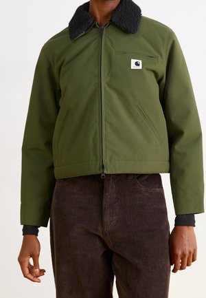 Personne portant une veste zippée verte avec un col en polaire noire et un pantalon en velours côtelé marron, debout devant un fond uni.