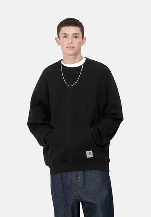 Sudadera negra con cuello redondo, mangas largas, bolsillos frontales y un pequeño parche de logo. Usada sobre una camiseta blanca con jeans azules.