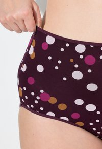 Eine Hand zieht den Bund von lila Unterwäsche mit weißen, rosa und braunen Punkten an einer hellhäutigen Hüfte.