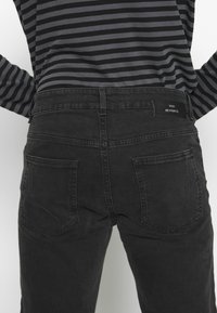 Los jeans de denim negro presentan un diseño clásico con dos bolsillos traseros y una etiqueta de cuero. La tela tiene una textura sutil y un acabado desteñido.