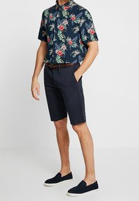 Chemise à manches courtes bleu marine avec motifs floraux rouges et verts tropicaux, associée à un short foncé et des chaussures foncées à enfiler. Ceinture marron incluse.