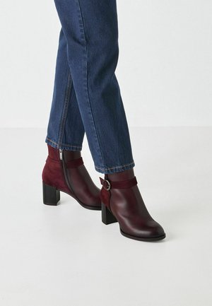 Bottines bordeaux à finition mate, avec un talon épais, des accents en daim et une fermeture éclair sur le côté. Associées à un jean bleu foncé.