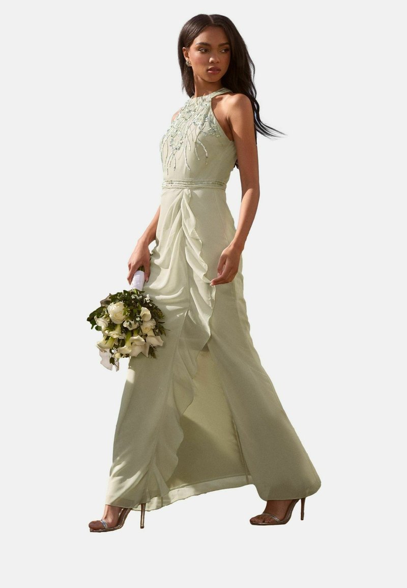 Helles grünes Kleid mit Halterneck, verziertem Oberteil, Rüschenshirt und fließender Silhouette. Das Model hält einen Strauß aus weißen Blumen.