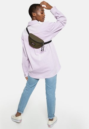 Sac banane vert olive avec fermeture éclair, attaché à une sangle noire, porté sur une chemise oversize violette claire et un jean bleu clair. Baskets décontractées.