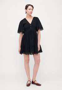 Schwarzes Kleid mit kurzen Rüscheärmeln, einem V-Ausschnitt und dekorativen Knöpfen. Mit Lochmusterbesatz und einem gestuften Rock. Glatte Textur.